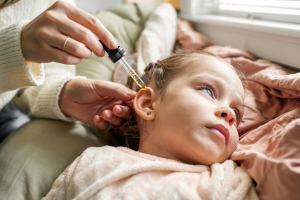 A young child lies on her side on a cozy bed while an adult gently gives ear drops with a glass dropper, illustrating a tender moment of using home remedies for eardrum pain.